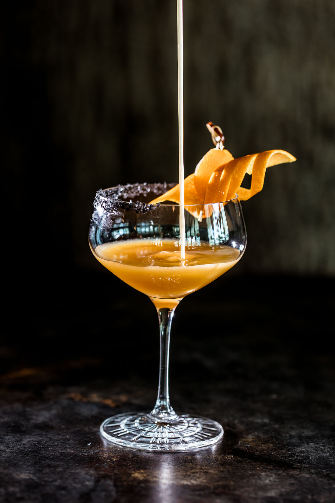 Orange cocktail being poured into fancy coup glass with exotic garnish