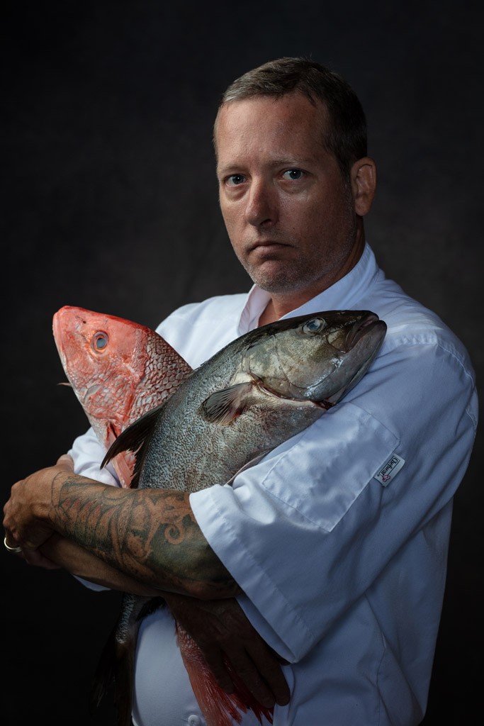 Intense photo of chef with tattoos holding two raw fish to his chest