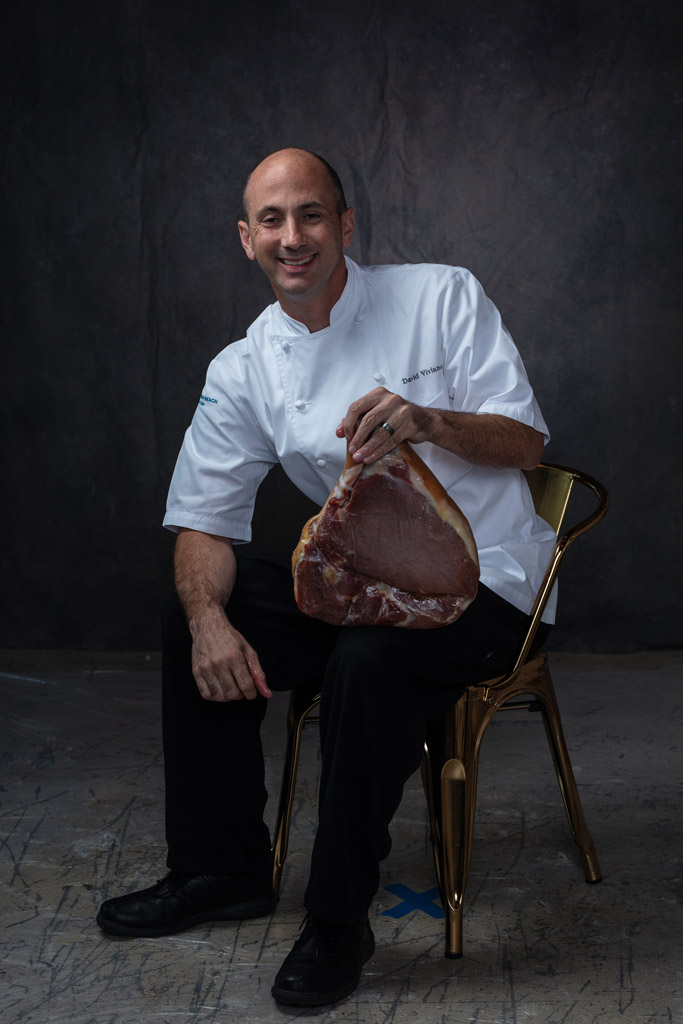 Bald chef sitting and holding large cut of raw meat with big smile