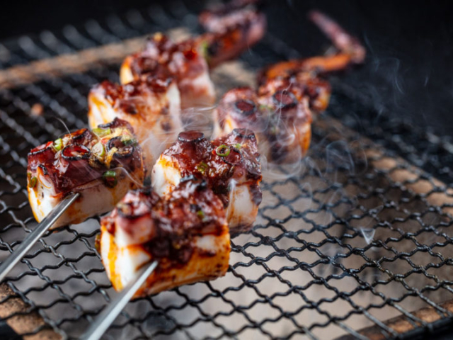 Octopus skewers cooking over a tabletop grill with smoke