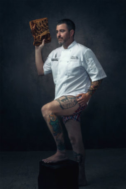 Hot chef without his pants on and covered in tattoos holds meat.