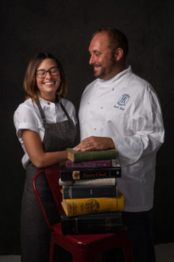 Chef and pastry chef who are married pose behind a stack of cookbooks